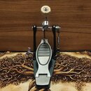 Černý basový pedál s kovovým šlapadlem a stříbrnými detaily. Horní část obsahuje mechanismus s řetězem, kovovou tyčou a bílým plstěným tlumičem. Povr…