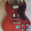 Červená elektrická kytara s černým pickguardem a dvěma humbuckery. Na těle je nalepený modrý kulatý štítek s nápisem ODVAHAVA VOLIT. Nástroj má čtyři…