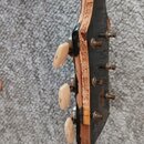 Detail hlavy kytary s černým povrchem a světlými ladicími kolíky. Kolem okrajů hlavy je viditelné výrazné opotřebení a odlupávání laku i materiálu. N…
