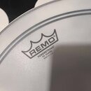 Detailní záběr na bílou, potaženou bicí koženu. Na povrchu je černý nápis REMO s logem koruny a text POWERSTROKE® P3 COATED MADE IN U.S.A. Kožena je…