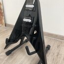 Černá elektrická kytara s ostrým, trojúhelníkovým tvarem těla umístěná na černém stojanu. Nástroj má dva humbuckery s bílými plastovými krytkami, na…