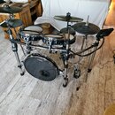 Elektronická bicí souprava s černými pady a černými činely na stříbrném kovovém rámu. Na basovém bubnu je nápis „ROLAND“ a „V-Drums“. Souprava obsahu…