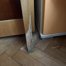 Detail dřevěné nohy nábytků v přirozené barvě s kovovou patkou na spodní části. Spodní část nohy vykazuje výrazné opotřebení, odloupaný lak a praskli…