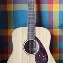 Akustická kytara s přírodním světlým povrchem korpusu a tmavým hmatníkem. Na hlavě je nápis YAMAHA a logo znázornující tři vlnovky. Kytara má tmavě h…