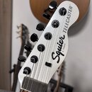 Detail hlavy elektrické kytary v bílé barvě. Na povrchu je černý nápis Squier by FENDER TELECASTER. Kytara má šest černých ladicích kol, strmý tvar h…