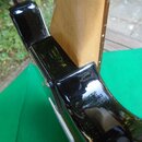 Detailní pohled na spoj hnedebního nástroje, pravděpodobně kytary, kde je světlý dřevěný krk zasazen do černého lakovaného korpusu. Černý lak vykazuj…