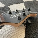 Detail hlavy kytary v matně černé barvě s viditelným dřevěným jádrem na hranách. Na povrchu je bílý nápis Sterling by MUSIC MAN a bílý podpis. Jsou v…