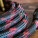Detailní záběr na svinutý kabel v textilním opletení. Kabel má černou barvu s červenými vzory a je zakončen černým plastovým konektorem. Povrch oplet…