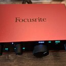 Červený kovový audio rozhraní s nápisem Focusrite na horní straně. Přední panel obsahuje čtyři černé ovládací knoflíky, několik malých modrých kontro…