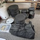 Sada černých polstrovaných obalů na bubny různých velikostí a jeden velký obdélníkový přepravní vak. Vedle obalů je položen buben s nápisem TAMA na b…