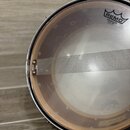 Fotografie znázorňuje snare drum z pohledu shora. Na průhledném koženě je vidět logo REMO a nápis AMBASSADOR® SNARE SIZE MADE IN U.S.A. Přes kožen pr…