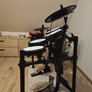 Elektronická bicí souprava na černém kovovém stojanu s několika padovými plochami bílé barvy. Na pedálech je viditelný nápis Alesis. Povrchy padů a k…