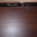 Detailní pohled na horní hranu hnědého dřevěného povrchu s černými štítky. Na štítcích jsou bílé nápisy HAMMOND EXP-100, BASS TREBLE TONE CONTROL, R…