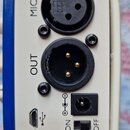 Detail zadní strany modrého zařízení s krémovým předním panelem. Jsou zde vidět dva XLR konektory s nápisy MIC a OUT, USB port s ikonou, napájecí vst…