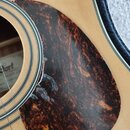 Detailní záběr na spodní část ozvukové otvory akustické kytary se světlým dřevěným povrchem. Na těle je nalepený hnědý pickguard s tmavou žilnatou st…