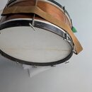 Dřevěný buben s hnědým povrchem a černými obručemi. Má bílou membránu a je vybaven hnědým látkovým popruhem. Na povrchu dřeva a černých obručí jsou v…