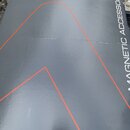 Detailní záběr na šedý povrch s nápisem MAGNETIC ACCESSORIES. Na povrchu jsou vidět tenké červené linky tvořící geometrické tvary. Materiál vykazuje…