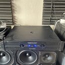 Černý výkonový zesilovač s nápisem AMERICAN AUDIO PROFESSIONAL POWER AMPLIFIER a označením modelu VLP1500. Přední panel obsahuje dva ovládací knofíky…