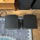 Dvě černé, kvádrové elektronické jednotky s žebrovaným chladičem na horní straně, umístěné vedle sebe na šedém koberci. Povrchy jsou matně černé a vy…