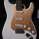 Bílá elektrická kytara typu Stratocaster s hnědým pickguardem z broušeného kovu. Nástroj má tři bílé snímače, tři ovládací knofíky s nápisy VOLUME a…
