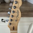Fotografie zachycuje hlavu elektrické kytary z přírodního světlého dřeva s chromovanými ladičkami. Na povrchu je černý nápis Fender® a pod ním menší…