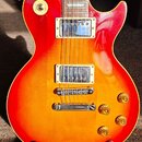 Elektrická kytara s trupem v červeno-žlutém sunburst provedení a hnědým hmatníkem. Nástroj má dva humbuckery s kovovými kryty, stopku typu Tune-o-mat…