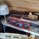 Červený přenosný radiomagnetofon s nápisy AIWA a CS-230. Zařízení má dvě velké černé reproduktory a stříbrné ovládací prvky. Je umístěno na bílé poli…