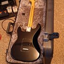 Černá elektrická kytara s tvarem Stratocaster a světlým dřevěným hnedem krku leží v otevřeném tvrdém kufru s šedým polstrováním. Na kytare není vidět…