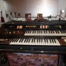 Černý dvoumanuálový elektronický orgán s bílými a černými klávesami. Nad horním manuálem jsou umístěny řady barevných přepínačů a ovládacích prvků. N…