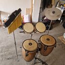 Sada perkusních nástrojů tvořená čtyřmi kongami z dřeva s světlými membránami a kovovýmivypnutými mechanismy. Vedle nich stojí na stojanu žlutý kovov…