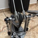 Fotografie zachycuje detail mechanismu basového pedálu v kovovém provedení. Je vidět řetězový pohon s ozubeným kolem a dvě kovové tyče. Povrch vykazu…