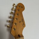 Záběr na hlavu elektrické kytary v zlatavé barvě s viditnou strukturou dřeva. Na zadní straně je černý nápis Fender STRATOCASTER a menší text "WITH S…