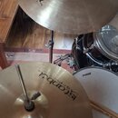 Fotografie zachycuje část bicí sady, konkrétně dva mosazné činely a černý buben. Na horním činely je vidět logo „Istanbul Agop Turkey“ a nápis „XIST“…