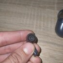 Fotografie zachycuje jedno bezdrátové sluchátko v tmavě šedé barvě, které drží ruka. Na spodní části sluchátka je viditelné bílé písmeno R. V pozadí…