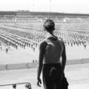 Na fotografii není žádný hudební nástroj ani příslušenství. Snímek zobrazuje muže v tílku zády k fotografu, který se dívá na rozsáhlý shromážděný dav…