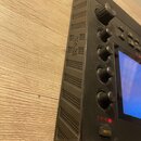 Černý elektronický přístroj s plastovým povrchem a řadou pěti ovratových ovládacích knoflíků. Na boku je vyraženo logo NOVATION a v dolní části se na…