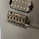 Detail elektrické kytary v šedé barvě. Je vidět černý humbucker a černý most s nápisem Sterling. K mostu je připojen chromovaný pákový systém. Povrch…