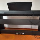Černý digitální piano v černém stojanu s přikryvním krytem kláves. Na levé straně horního panelu je bílý nápis YAMAHA s logem značky. Z přístupového…