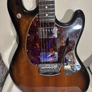 Elektrická kytara s tmele s hnědým sunburst povrchem a hnědým hmatníkem s bílými tečkovanými značkami. Nástroj má tmavě červený želvový pickguard, je…