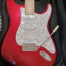 Červená elektrická kytara typu Stratocaster s bílo-perleťovým pickguardem, umístěná v černém obalu. Nástroj má javorový hmatník a tři snímače s bílým…