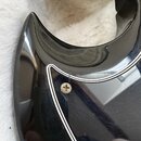 Detail černé lakované části těla elektrické kytary s bílým lemováním. Povrch vykazuje drobné škrábance a v místě u šroubu je viditelná prasklina v la…