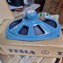 Modrý reproduktor s kovovým krytem a středovým knoflíkem, na kterém je nápis TESLA. Reproduktor leží na hnědé kartonové krabici s velkým modrým nápis…