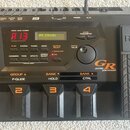 Šedý kytarový syntetizátor Roland GR-33 v plastovém provedení. Na horním panelu je digitální displej s nápisem A13 JP8 STRINGS a různé ovládací tlačí…