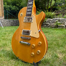 Elektrická kytara s žlutohnatým povrchem typu sunburst a krčkem z tmavého dřeva. Na hmatníku jsou vidět vložky s nápisem "Gibson" a na snímačích je n…