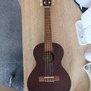 Tmavě hnědá ukulele s dřevěným korpusem a hnedým hmatníkem. Nástroj má čtyři struny, světlé ladící kolíky a dřevěný kobylník. Uvnitř ozvučnice je vid…