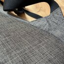 Detailní záběr na šedý textilní materiál s výraznou strukturou tkaniny. V horní části je vidět černý popruh z nylonu. Povrch materiálu vypadá čistě a…