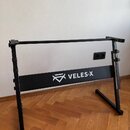 Černý kovový stojan na klávesové nástroje s hranatým profilem. Na přední vodorovné tyči je bílý nápis VELES-X doprovázený logem ve tvaru stylizovanéh…