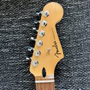 Záběr na hlavu elektrické kytary z přírodního světlého dřeva. Na povrchu je černý nápis Fender a STRATOCASTER. Jsou vidět šest chromovaných ladících…