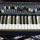 Černý syntetizátor s červenými bočními panely a bílo-černou klaviaturou. V pravém horním rohu je nápis Legend One ANALOG SYNTHESIZER. Horní panel obs…