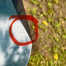 Detailní záběr na tmavě modrý, kovový povrch s jemnou texturou. V červeném kruhu je vidět odřápnutý lak a světlé poškození povrchu u okraje. Pozadí t…